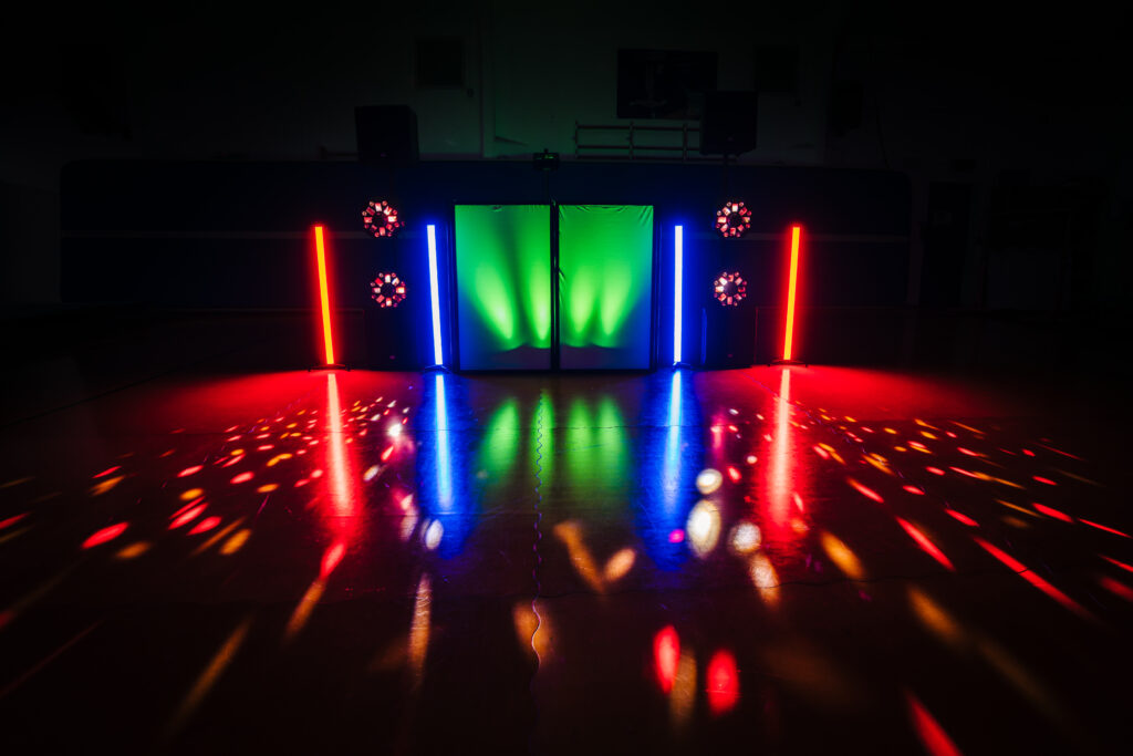 A dark room with vibrant lights reflecting on the floor. The setup includes vertical blue, red, and green lights, with colorful spotlights creating a dynamic and festive disco atmosphere. The reflections on the shiny floor add to the party vibe.